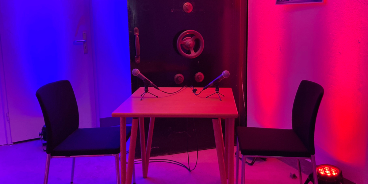 Neben der schweren Panzertür eines Tresors steht ein Tisch, daran zwei Stühle und darauf zwei Mikrofone. Scheinwerfer tauchen die Szene in Rot- und Blautöne.