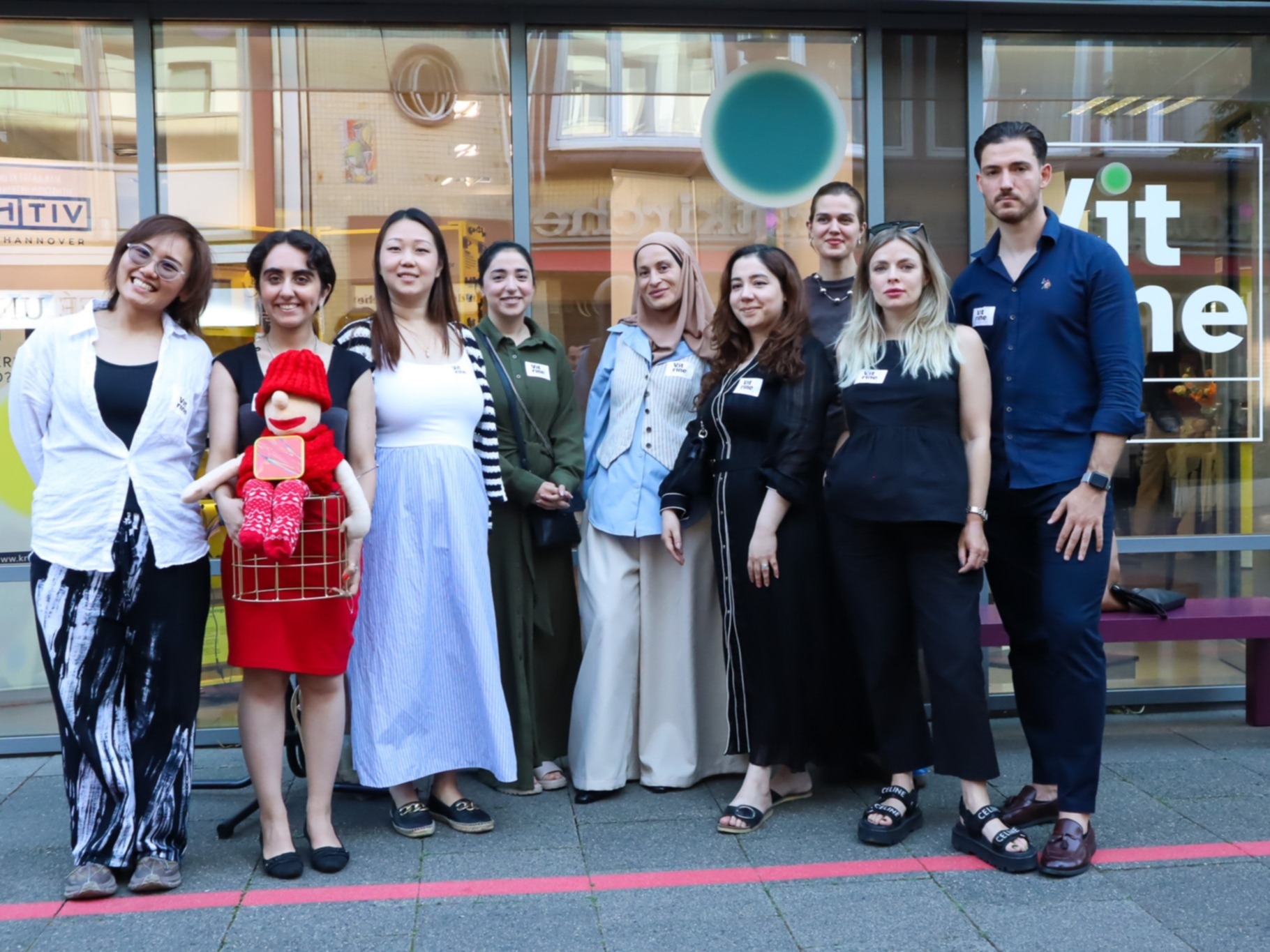 Neun Personen, die f&uuml;r ein Gruppenbild posieren. &Uuml;berwiegend Frauen. Alle stehen vor einer Schaufensterscheibe. Am Unteren Bildrand dre "Rote Faden", der durch Hannover f&uuml;hrt.