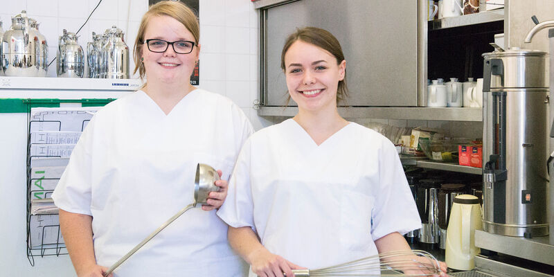 Zwei junge Frauen in weißer Berufskleidung stehen mit Kelle und Schneebesen in einer gewerblichen Küche.