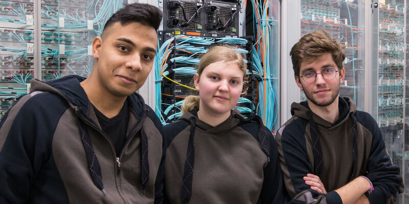 Zwei junge Männer und eine junge Frau stehen in einem Serverraum vor elektronischen Schaltschränken.