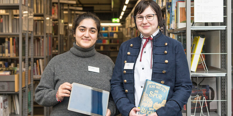 Zwei Auszubildende stehen zwischen Bücherregalen in der Stadtbibliothek; die eine hält ein Touchpad in der Hand, die andere ein Buch.